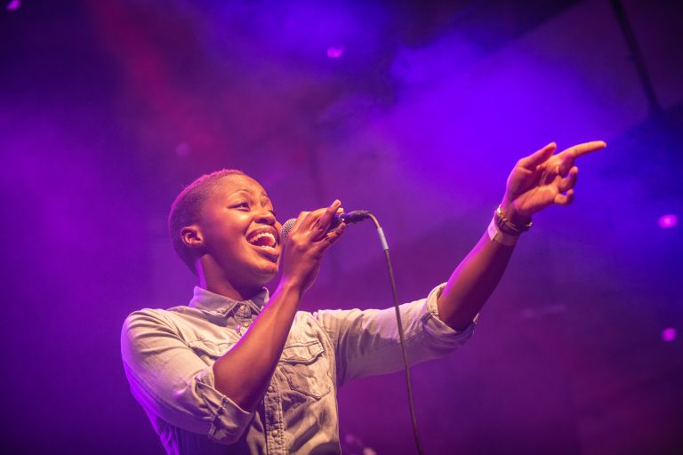 Een zanger met een microfoon staat op een podium, omringd door paarse verlichting.