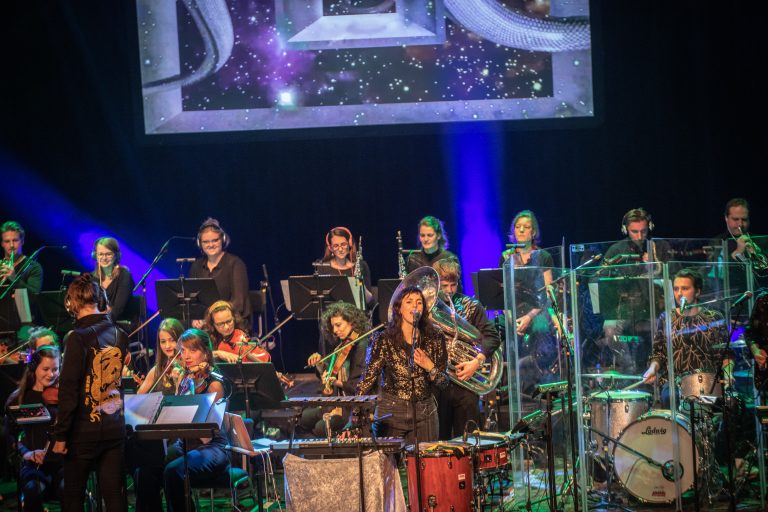 Een orkest met muzikanten op het podium, terwijl een soliste het publiek toespreekt.