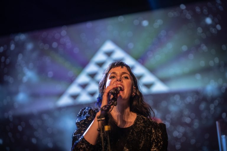 Een zangeres met microfoon, die optreedt tegen een kleurrijke visuele achtergrond.