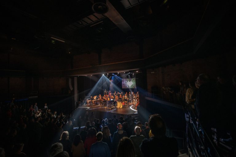 Een groep muzikanten optredend op een podium, verlicht door stralen licht, met een publiek dat toekijkt.