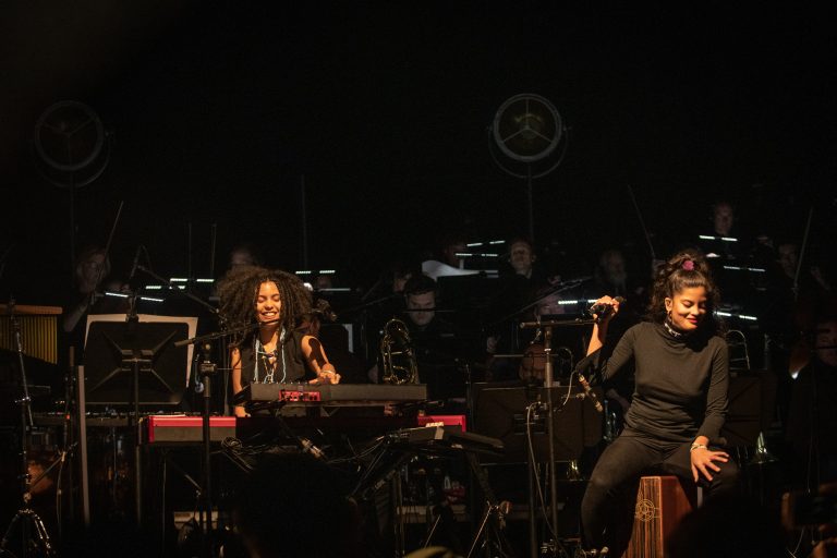 Twee vrouwelijke muzikanten optreden op een podium, met een keyboard en microfoon, omringd door een orkest.
