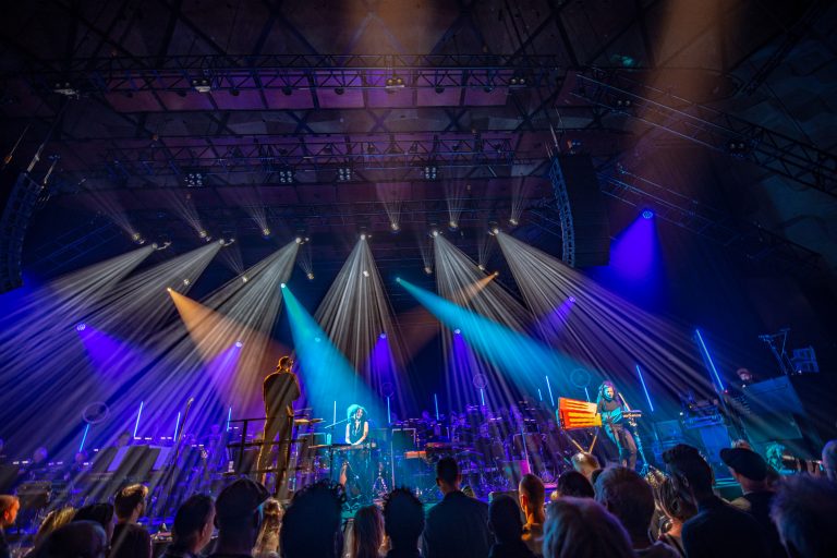 Een live concert met kleurrijke lichtstralen die over het podium schijnen, terwijl het publiek toekijkt.