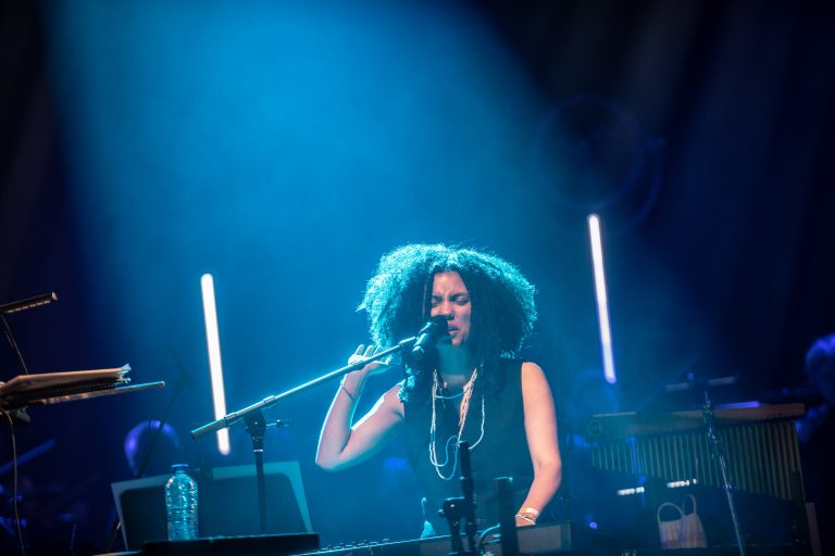 Een muzikante met een volle, krullende haardos zingt achter een piano, omringd door blauwe lichtstralen.