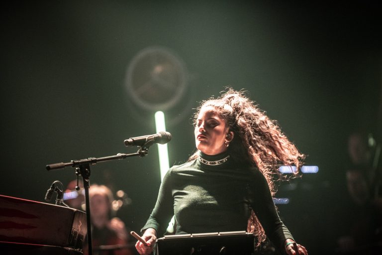 Een muzikante met lang, golvend haar zingt en speelt voor een publiek, terwijl sfeerverlichting op de achtergrond aanwezig is.