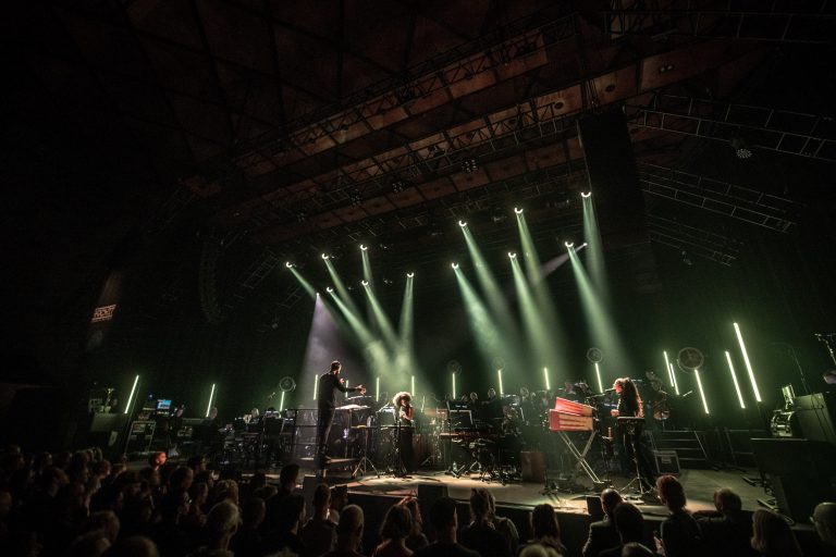 Een live muziekoptreden met meerdere artiesten op het podium, omringd door sfeervolle verlichting en een enthousiast publiek.