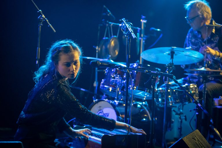 Een muzikant speelt op een keyboard met een drumstel op de achtergrond, verlicht door blauwe lichten.