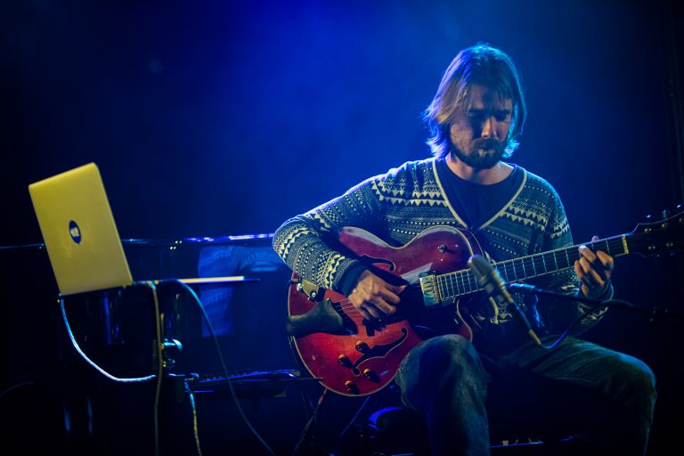 Een muzikant speelt een rode gitaar op een podium met blauwe verlichting.