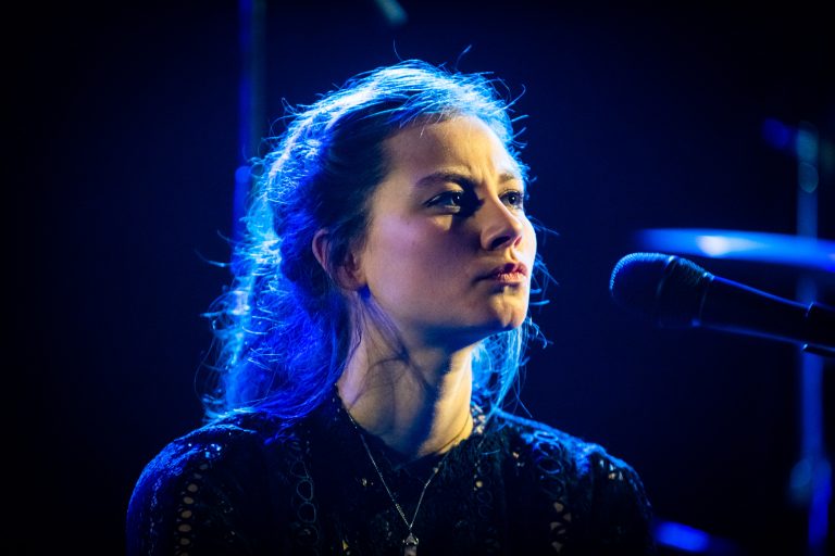 Een vrouw met een intense blik, verlicht door blauwe lichten, staat naast een microfoon.