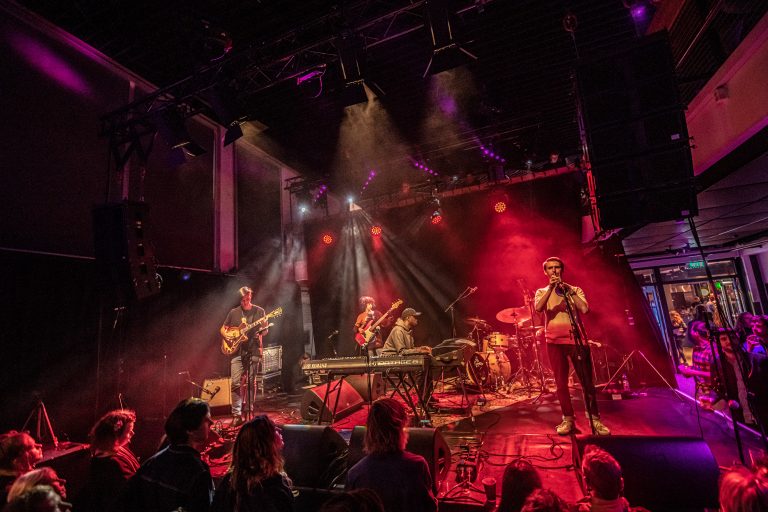 Een band speelt op het podium met kleurrijke lichten en een publiek in de voorgrond.