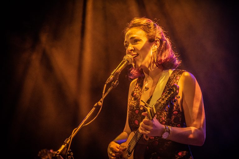 Een muzikante zingt en speelt op een podium, omringd door warme, gouden belichting.