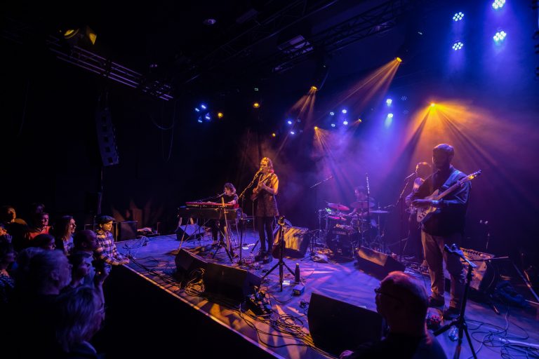 Een live muziekoptreden op een donker podium met kleurrijke verlichting en muzikanten die spelen.