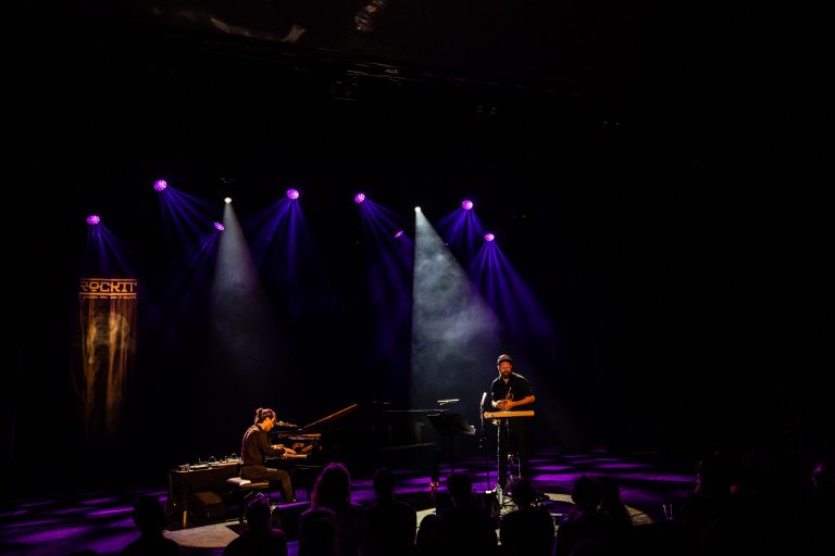Twee muzikanten optreden op een podium met paarse verlichting.