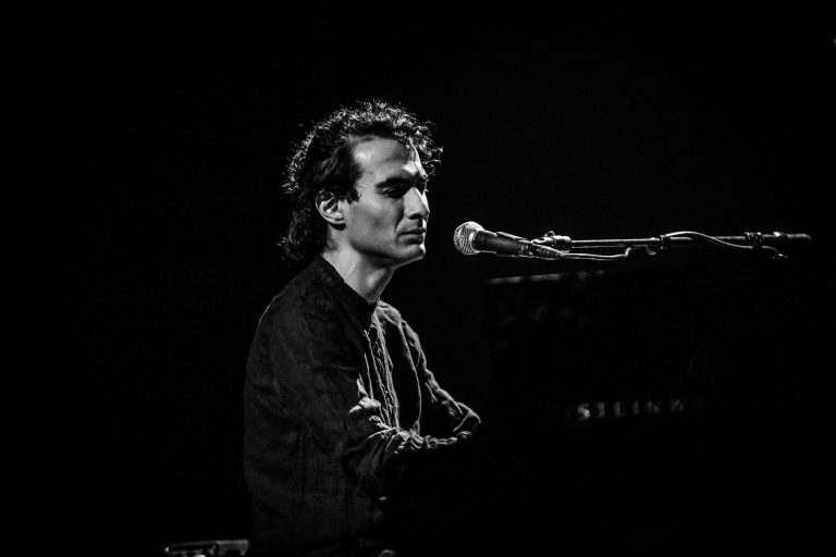 Een muzikant zittend achter een piano, terwijl hij in de microfoon zingt in een donkere setting.