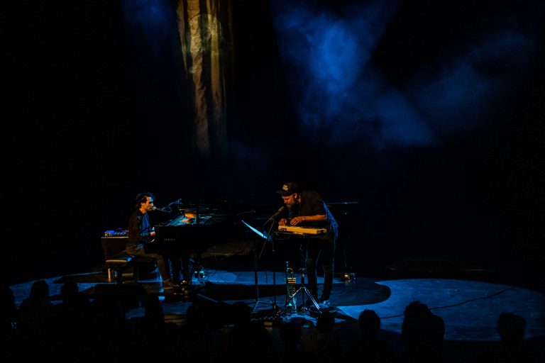 Twee muzikanten spelen piano op een donkere podium met blauwe belichting.