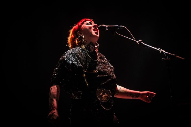 Een zangeres met een rood haar en opvallende kleding staat op het podium terwijl ze in de microfoon zingt.