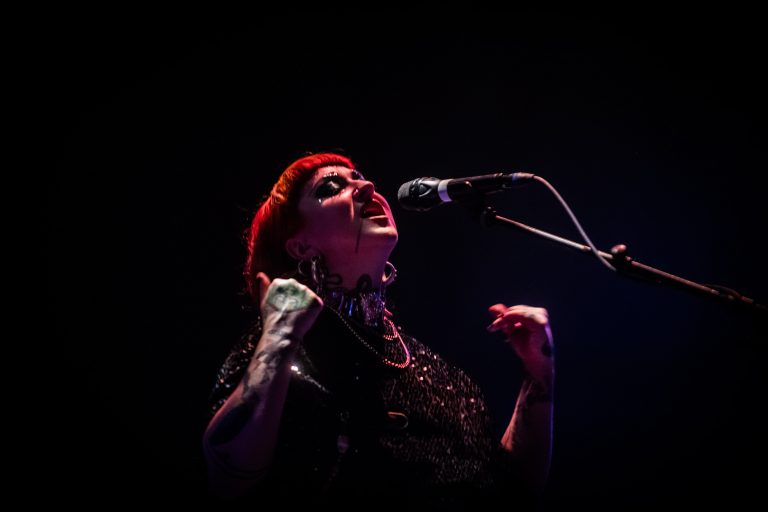 Een zangeres met rood haar zingt passievol in een donkere omgeving, met een microfoon voor haar.