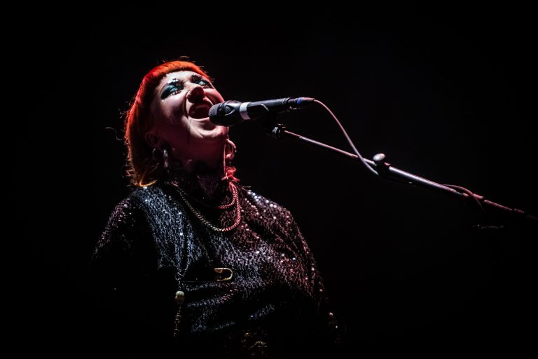 Een zangeres met rood haar die enthousiast in een microfoon zingt, verlicht door een enkele verlichting tegen een donkere achtergrond.