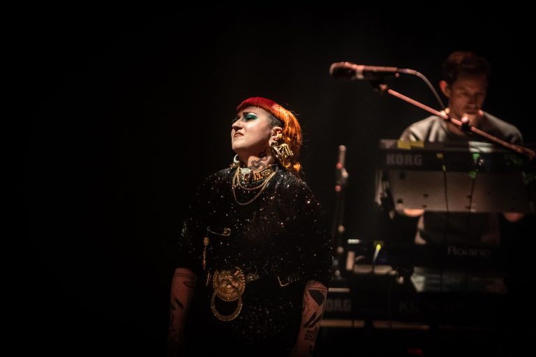 Een performer met een opvallende rode hoed en accessoires staat op het podium in een donkere omgeving.