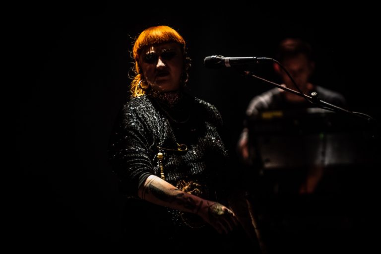 Een persoon met oranje haar staat op het podium met een microfoon in een sfeervolle, donkere setting.