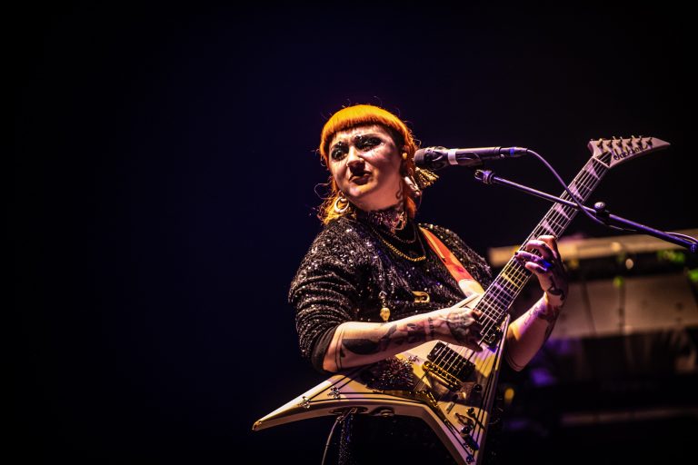 Een muzikant met een oranje hoofddeksel speelt gitaar en zingt in het licht van een podium.