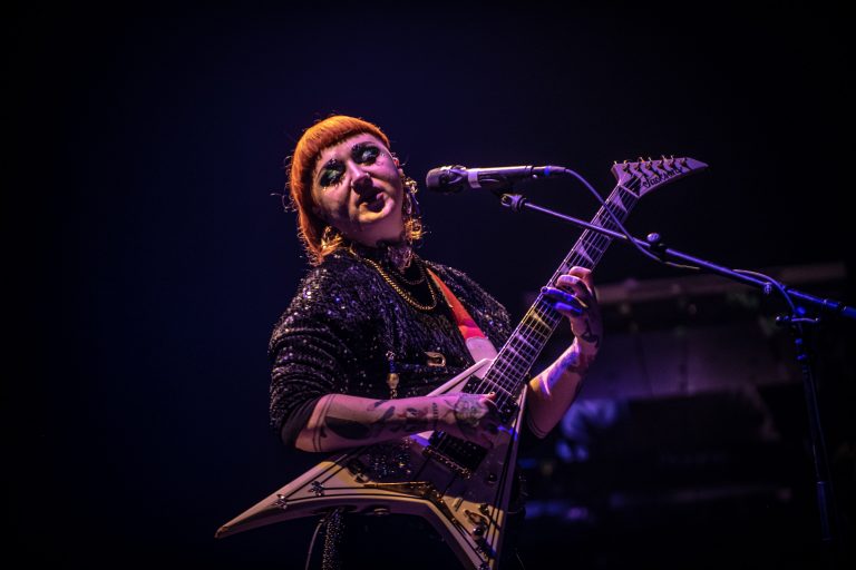 Een muzikante met een elektrische gitaar zingt op het podium in een donkere setting.