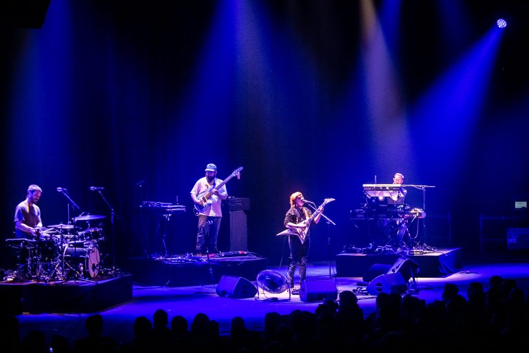 Een band optreden op een podium met blauwe verlichting, bestaande uit vier muzikanten met verschillende instrumenten.