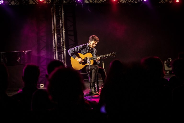 Een muzikant speelt gitaar op een podium met kleurrijke verlichting, terwijl publiek op de achtergrond kijkt.