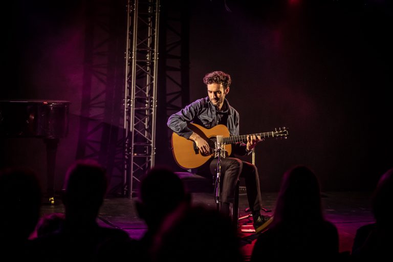 Een muzikant zit op een stoel en speelt gitaar voor een publiek in een sfeervol verlichte ruimte.