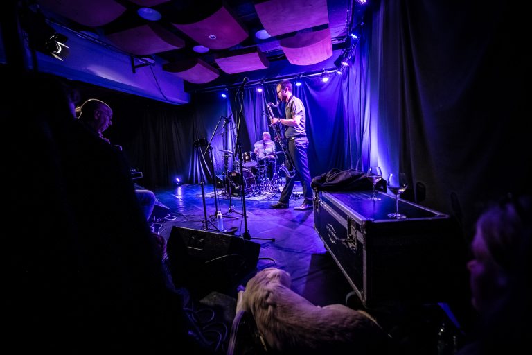 Een muzikant speelt op een podium met blauwe verlichting terwijl een drummer in de achtergrond zichtbaar is.
