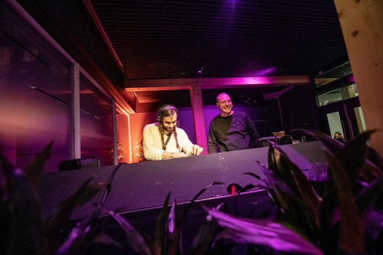 Twee DJ's staan achter een mengpaneel bij een evenement, verlicht door paarse en warme lichten.