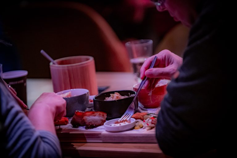 Een persoon eet van een bord met verschillende gerechten, met een focus op sushi en sauzen, te midden van een sfeervolle setting.