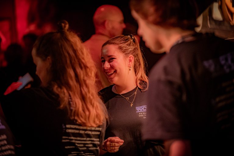 Een vrouw met een glimlach praat met anderen in een drukke, sfeervolle omgeving.