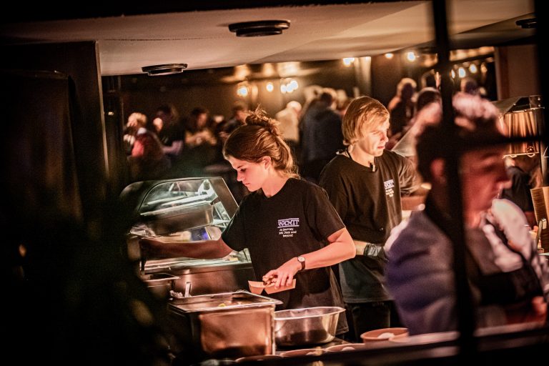 Twee medewerkers bedienen gerechten aan een buffet in een drukke eetomgeving.