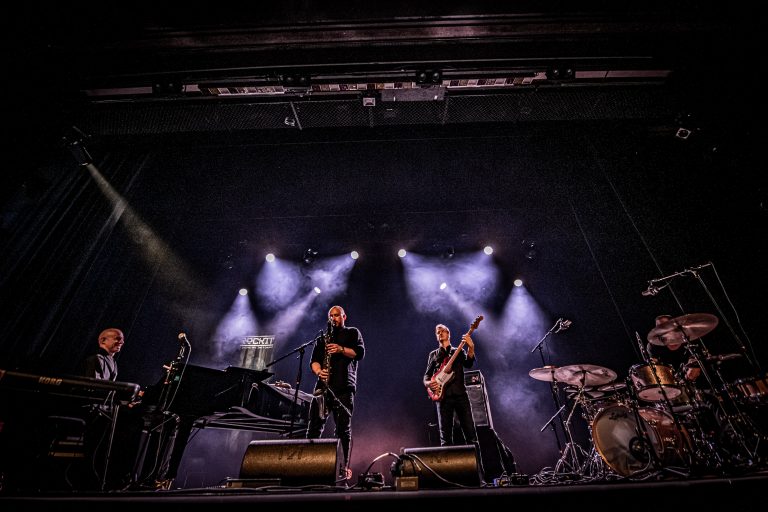 Een live band speelt op een podium met een piano, elektrische gitaar en drumstel, verlicht door meerdere lichten.