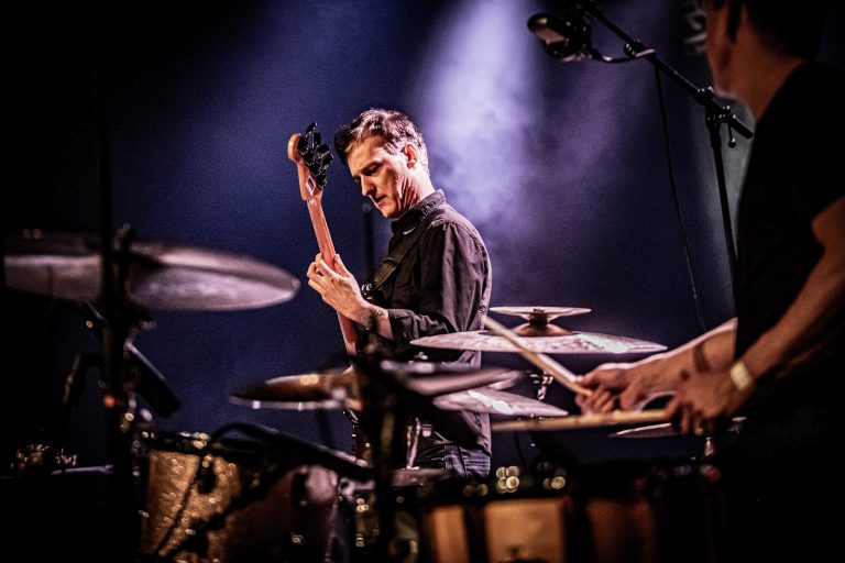 Een muzikant speelt op een elektrische gitaar tijdens een live optreden met een drums in de achtergrond.