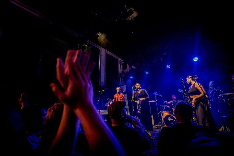 Een live muziekoptreden met een enthousiast publiek dat applaudisseert in een sfeervol verlichte zaal.