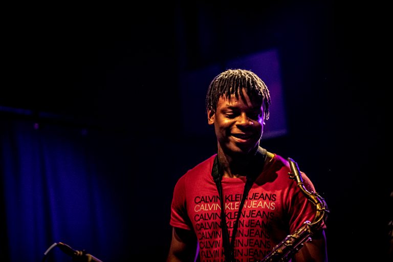Een muzikant met een saxofoon glimlacht terwijl hij op het podium staat, omringd door blauwe verlichting.