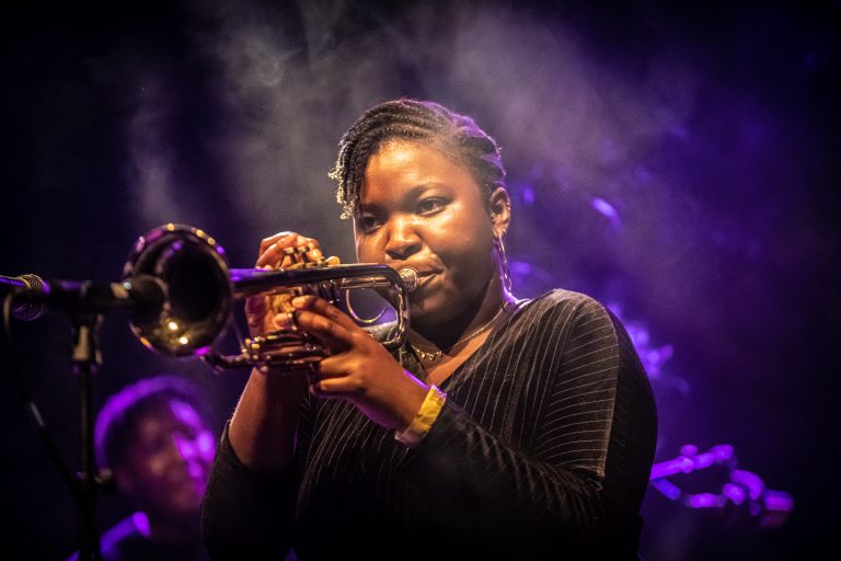 Een vrouw speelt trompet op een podium met een sfeervolle verlichting en rook in de achtergrond.