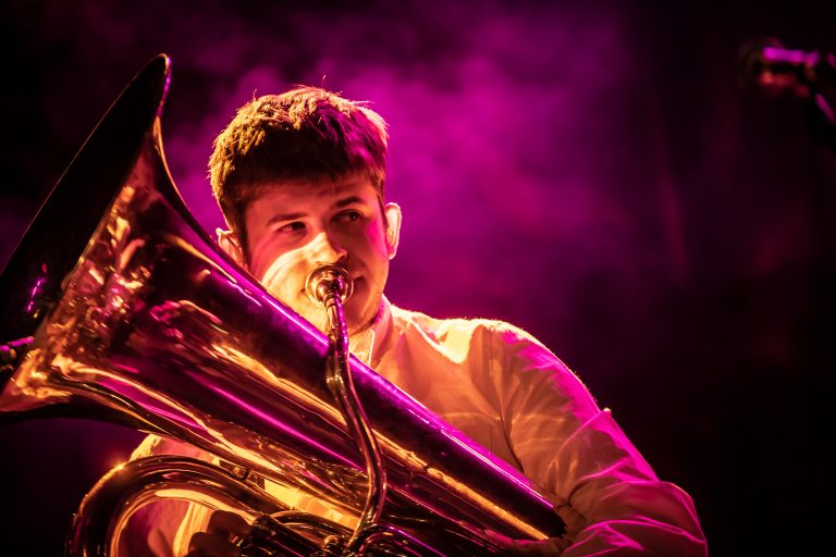 Een muzikant speelt een tuba op een podium, omringd door paarse rook.