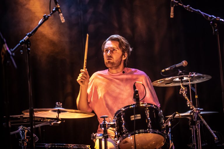Een drummer in een roze T-shirt speelt op een drumstel tijdens een optreden.