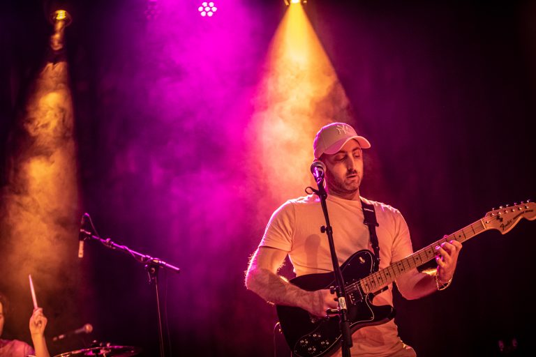 Een muzikant speelt gitaar op het podium met kleurrijke lichteffecten en een rookachtige achtergrond.