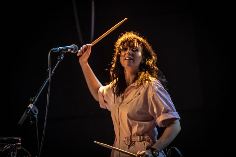 Een vrouwelijke drummer voert een energieke act op, terwijl ze stokjes vasthoudt en naar de camera kijkt.