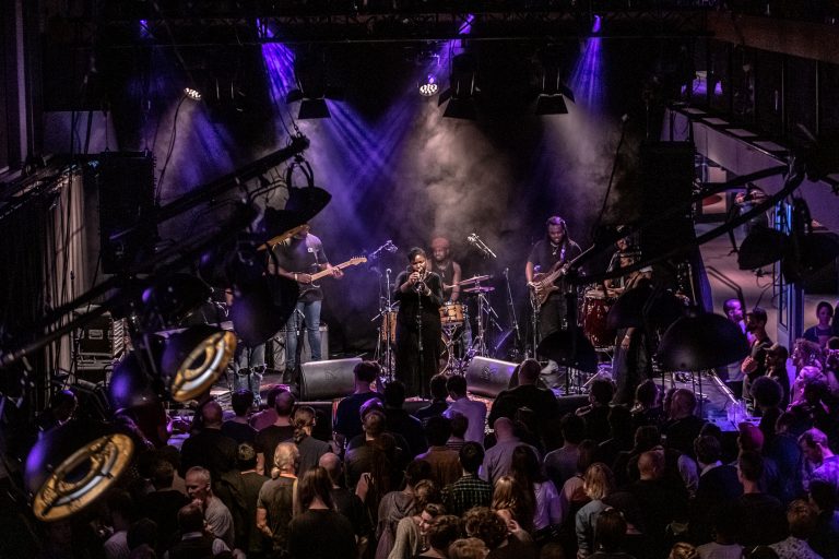 Een band optredend op een podium met een enthousiaste menigte en sfeervolle verlichting.