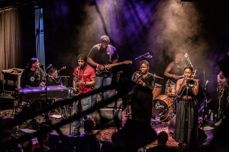 Een live muziekoptreden met een band bestaande uit verschillende muzikanten op een sfeervol verlichte podium.