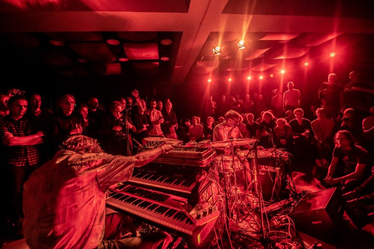 Een muzikant speelt op toetsenborden in een sfeervolle rode lichtsetting, omringd door een aandachtig publiek.