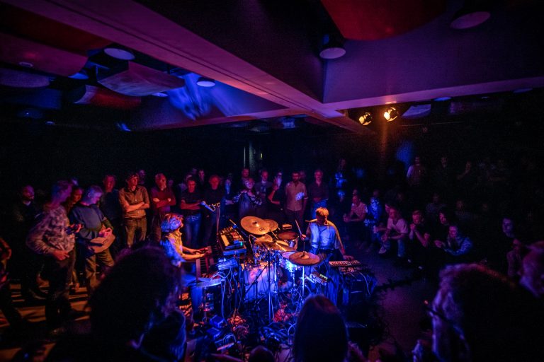 Een intieme muziekevenement met twee drummers op een podium omgeven door een aandachtig publiek in een sfeervolle verlichte ruimte.