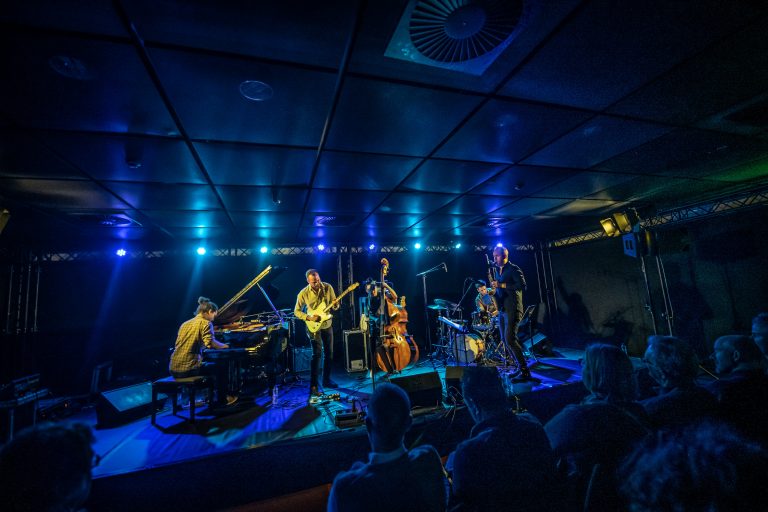 Een band speelt muziek op een podium verlicht met blauwe en groene lichten, terwijl het publiek toekijkt.
