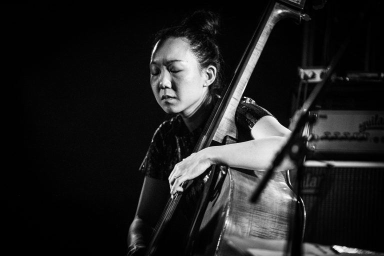 Een vrouwelijke muzikant speelt een contrabas in een donkere omgeving, terwijl ze zich concentreert op haar muziek.