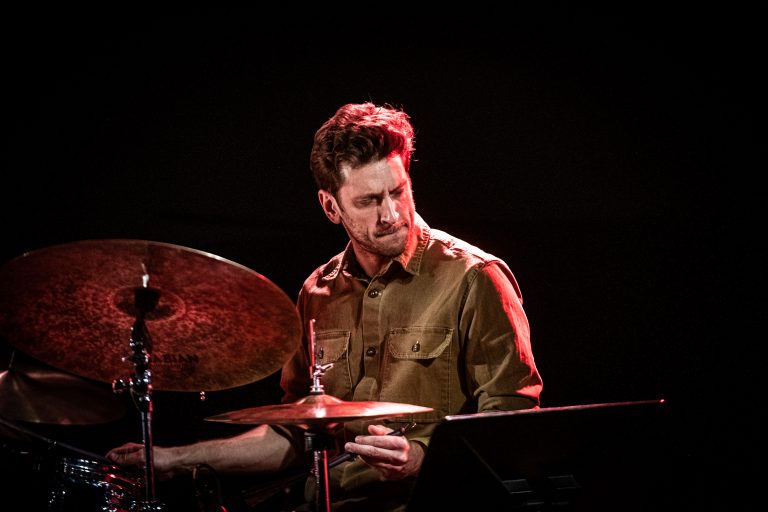 Een drummer in een bruin shirt speelt geconcentreerd op een houten drumstel, verlicht door een warme gloed.