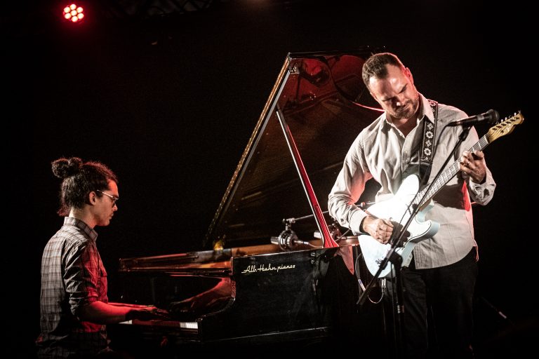 Twee muzikanten optreden op een podium, één speelt piano en de ander gitaar.